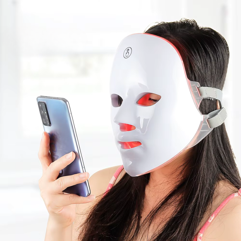 Woman wearing Luerelle Radiance LED Therapy Mask emitting red light while using a smartphone indoors